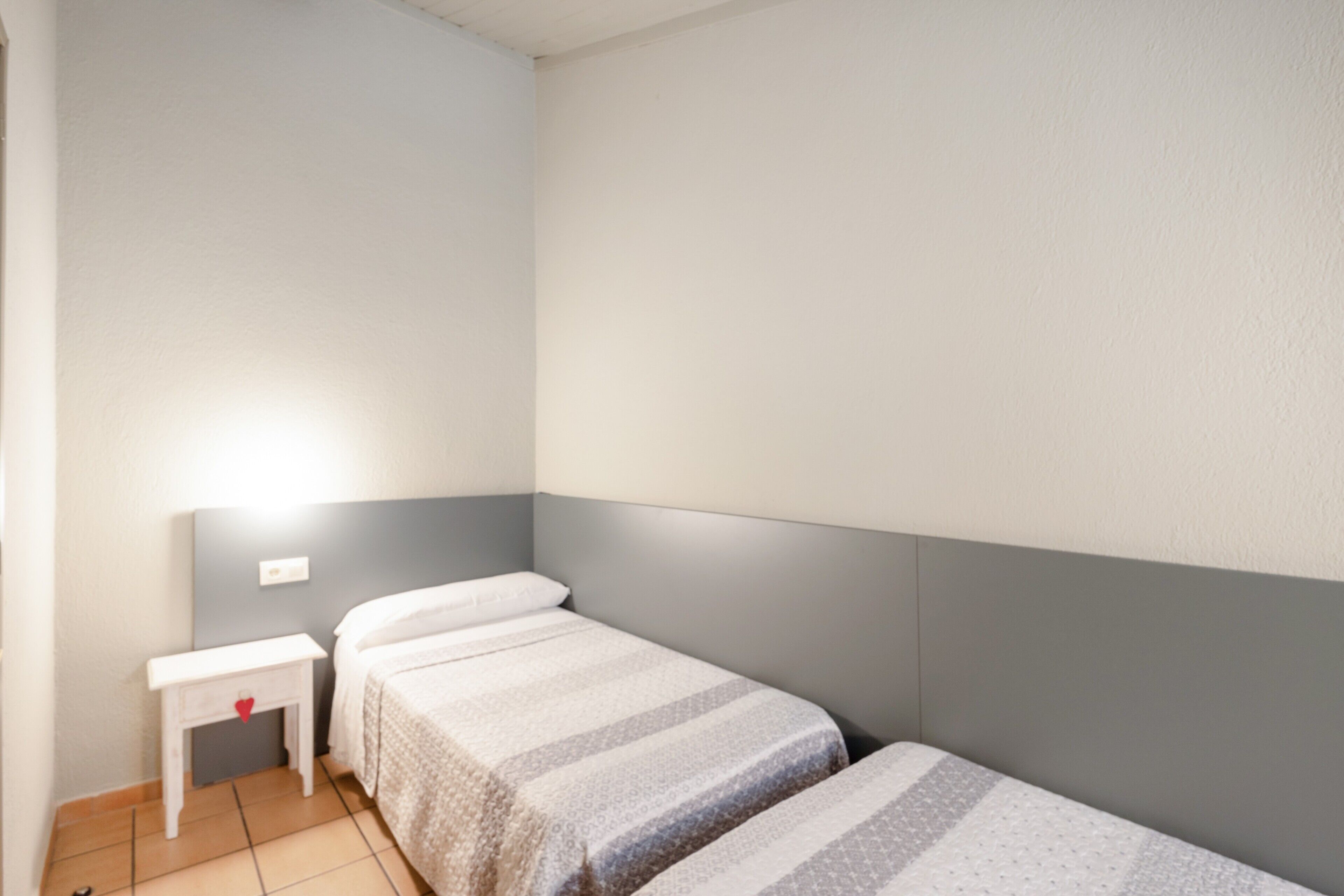 Une petite chambre avec un lit simple, une tête de lit grise, une table de chevet décorée d’un cœur rouge et une lampe fixée au mur.