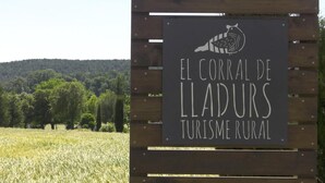 A sign with a bird illustration and text 'EL CORRAL DE LLADURS TURISME RURAL' against a backdrop of a field and trees. - Rural Apartment El Corral (Lladurs)