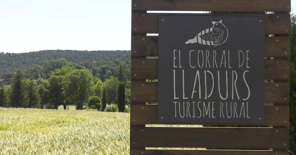 A sign with a bird illustration and text 'EL CORRAL DE LLADURS TURISME RURAL' against a backdrop of a field and trees. - Rural Apartment El Corral (Lladurs)