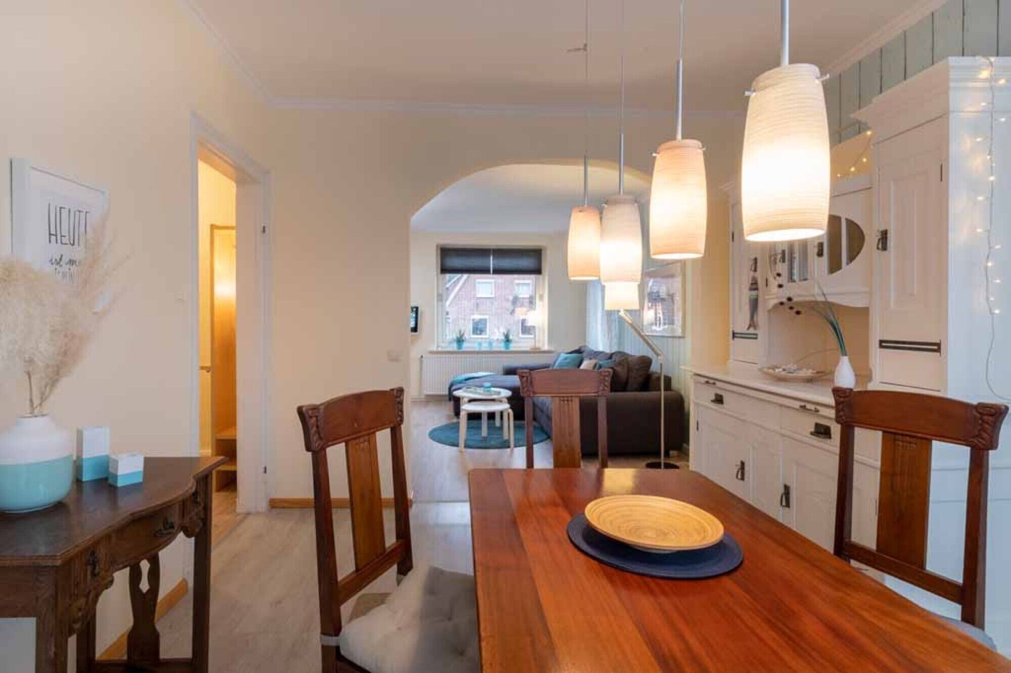 A dining area with a wooden table, chairs, pendant lights, and a view into the living room.
