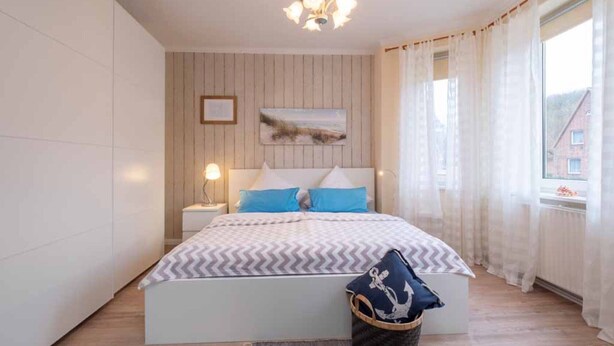 Un dormitorio con cama, mesita de noche, lámpara, cuadro en la pared, ventana con cortinas y vistas a edificios.