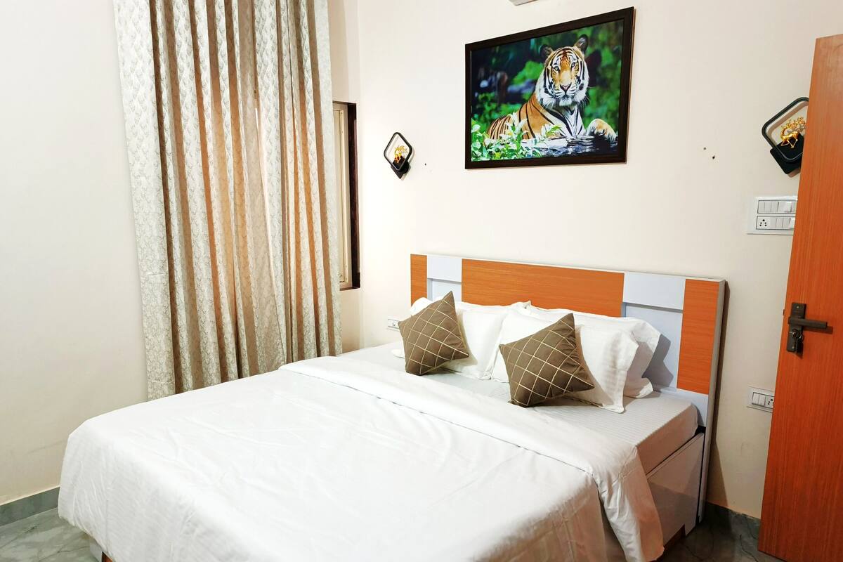 Una cama bien hecha con ropa de cama blanca y dos almohadas decorativas, un cuadro enmarcado de un tigre en la pared, y una puerta a la derecha.