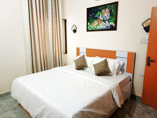 Uma cama bem arrumada com roupa de cama branca e duas almofadas decorativas, um quadro de uma tigresa na parede e uma porta de madeira à direita.