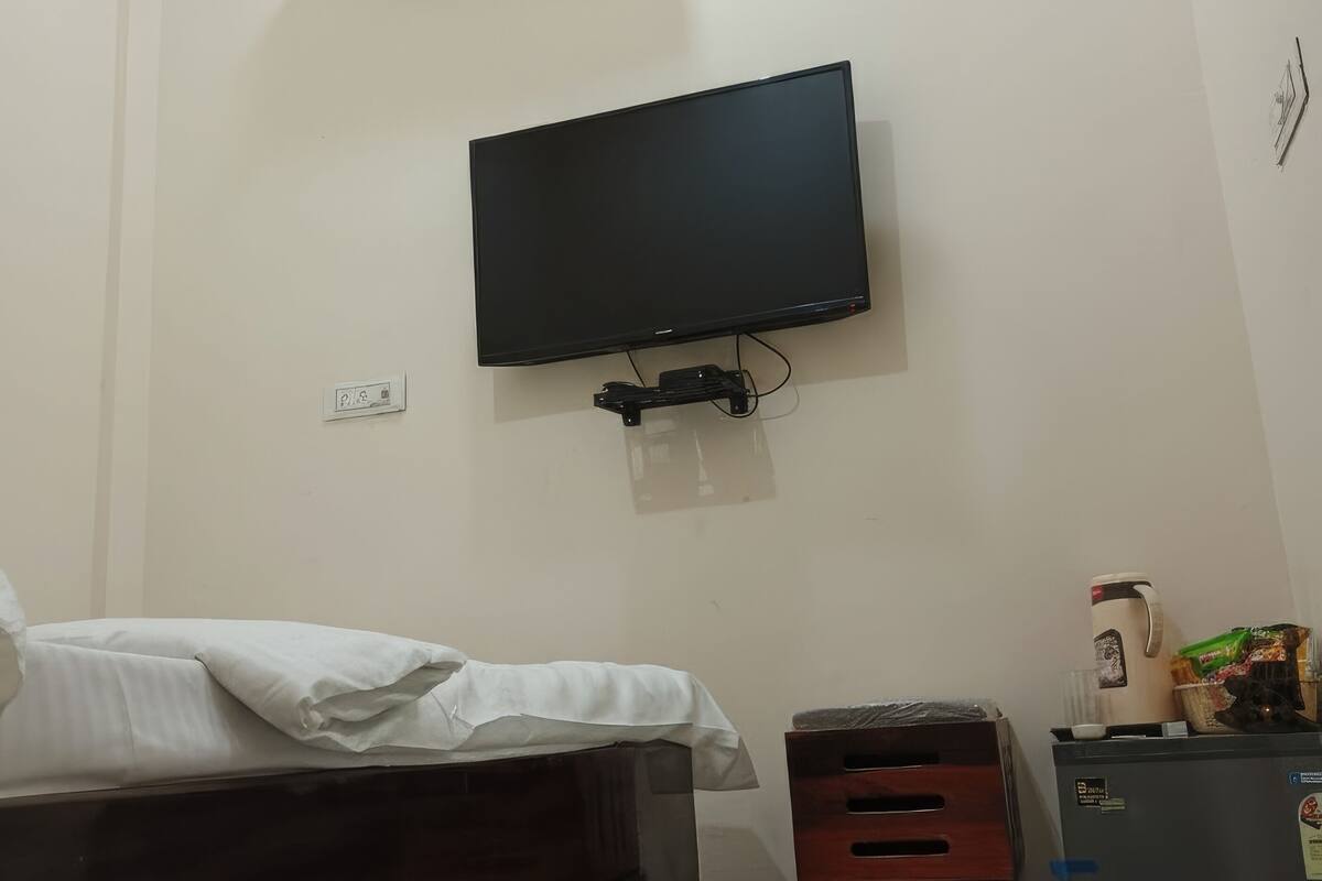 Un dormitorio con una cama, un televisor montado en la pared, aire acondicionado, un armario pequeño y un refrigerador.
