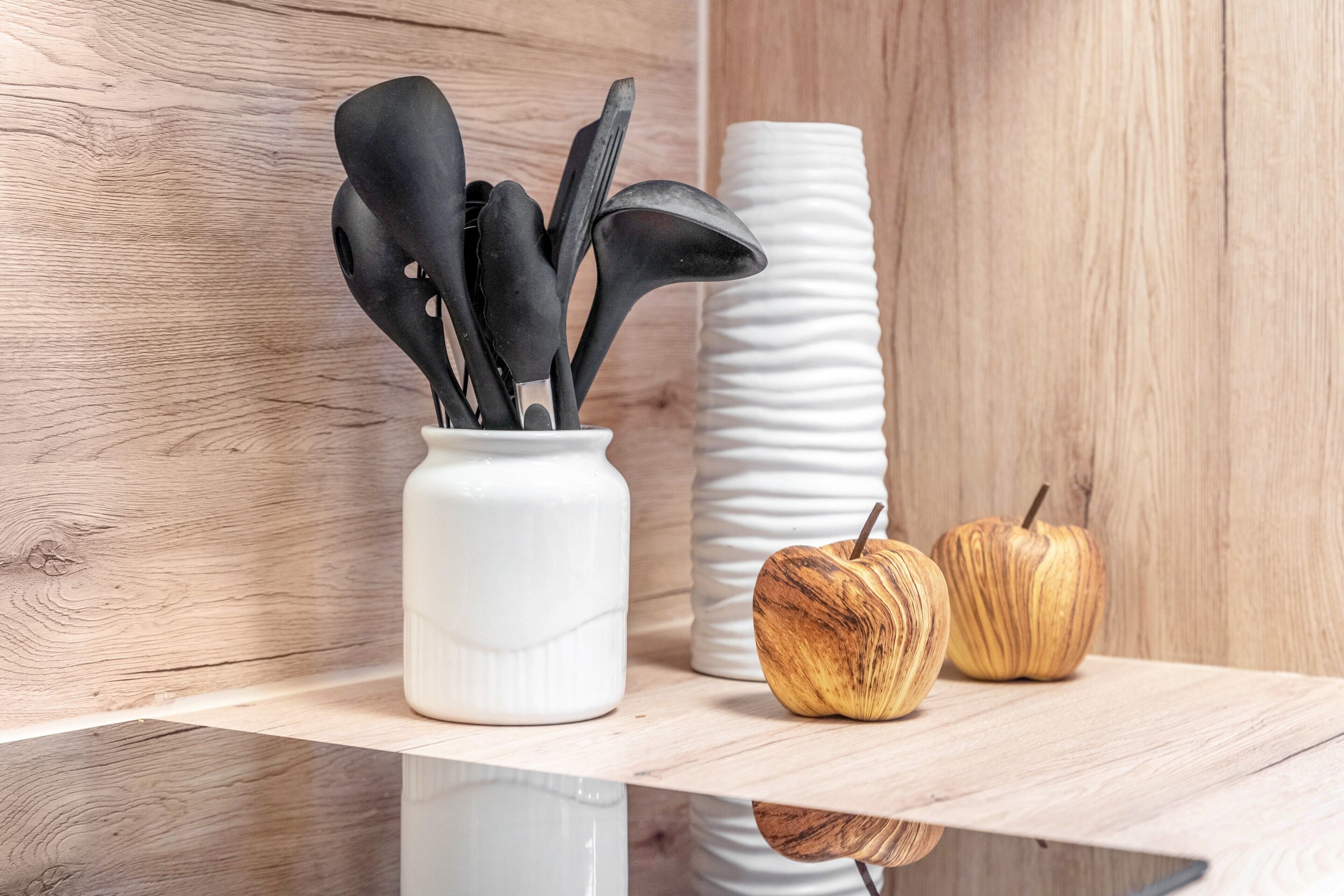 Eine weiße Keramikvase mit schwarzen Küchenutensilien und zwei hölzernen Äpfeln auf einem Holzregal.