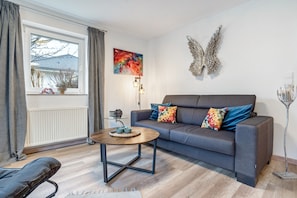 A living room with a grey sofa, a round coffee table, a window with curtains, a wall-mounted abstract painting, and a decorative winged wall art piece. - Apartment 'Carmen' with Shared Terrace, Private Garden and Wi-Fi (Wiek)