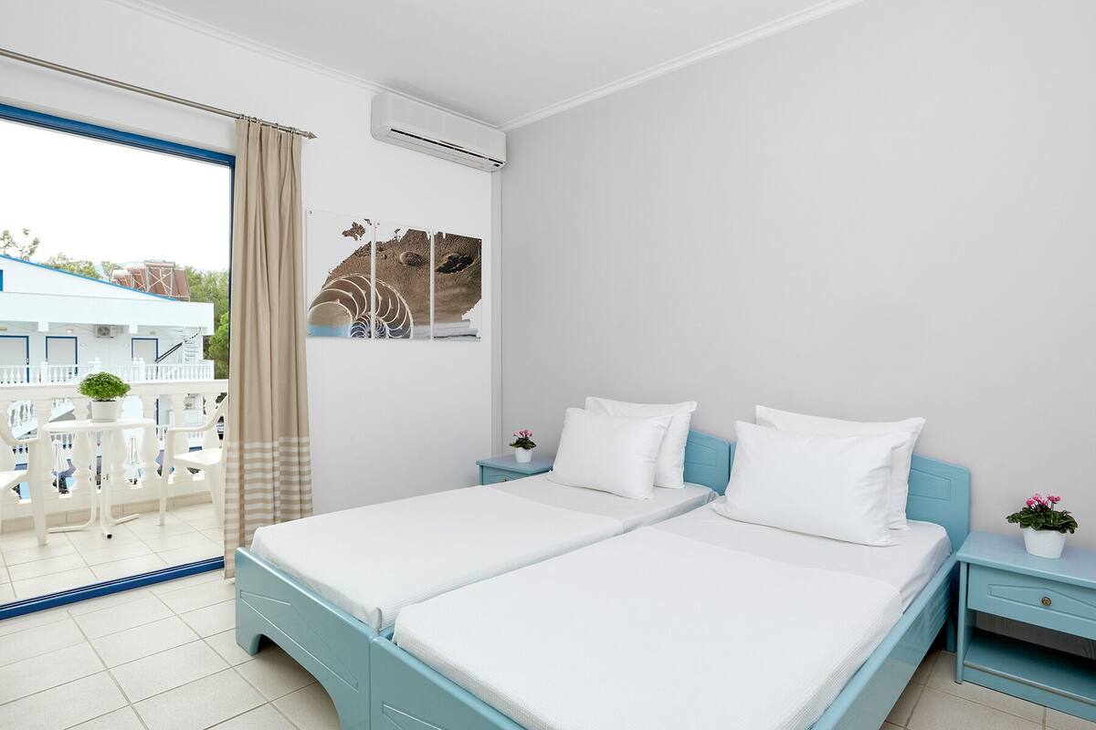 A hotel room with a bed, a small bedside table, a potted plant, and a view of the outside balcony.