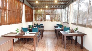 A restaurant with wooden tables and chairs, bamboo walls, and large windows. - Anandam Farms And Resort (Haridwar)