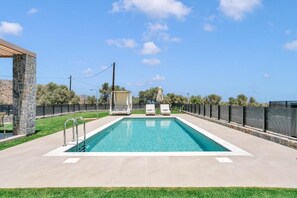 A rectangular swimming pool with a stone pillar on one side, two lounge chairs, and a pergola structure. - Olearia Villas (Malevizi)