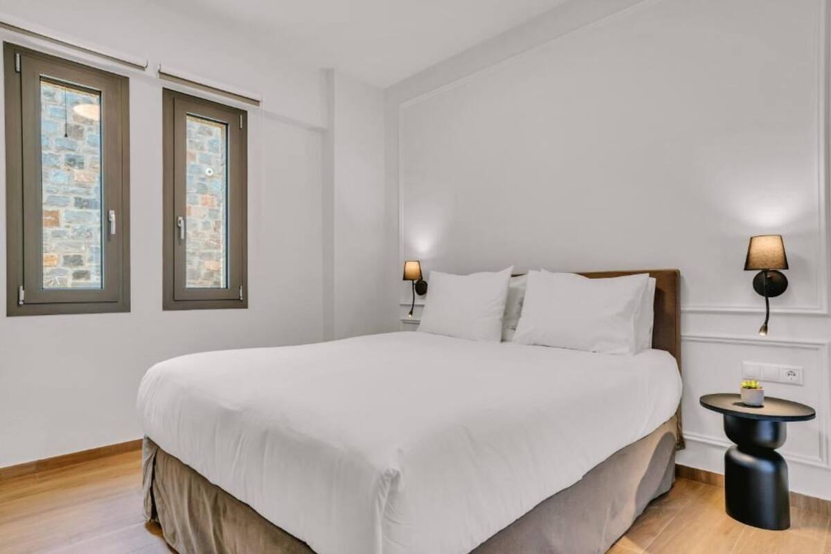 Une chambre avec un grand lit, deux fenêtres avec des encadrements en pierre, une table de chevet avec une lampe et un luminaire fixé au mur.