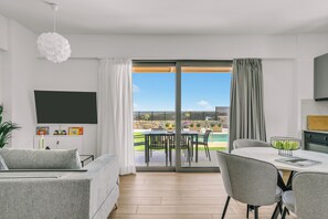 A modern living room with a sofa, a dining table with chairs, a TV mounted on the wall, and a large sliding glass door leading to an outdoor patio. - Olearia Villas (Malevizi)