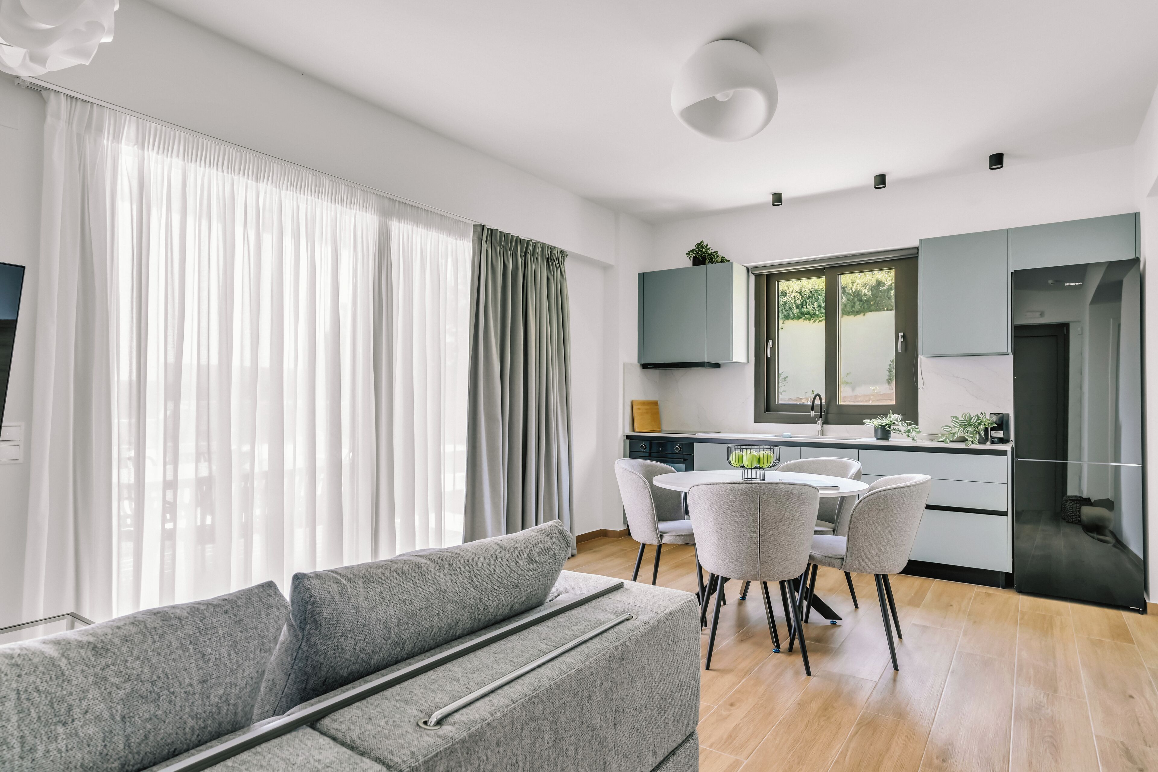 A modern living room with a grey sofa, a dining area with a round table and chairs, a kitchen with a black refrigerator, and a large window with sheer curtains.