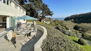 Un área de comedor exterior con curvas, con una mesa y sillas, una sombrilla azul y vistas al mar y las colinas.
