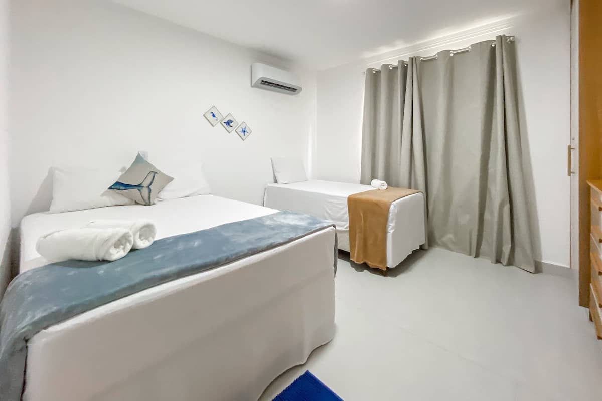 A hotel room with a bed, a bedside table, a small table, a curtain, and an air conditioning unit.