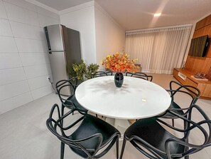 A dining area with a round table, black chairs, a vase with flowers, a refrigerator, and a sofa. - Residencial Saint Loius (Porto Seguro)