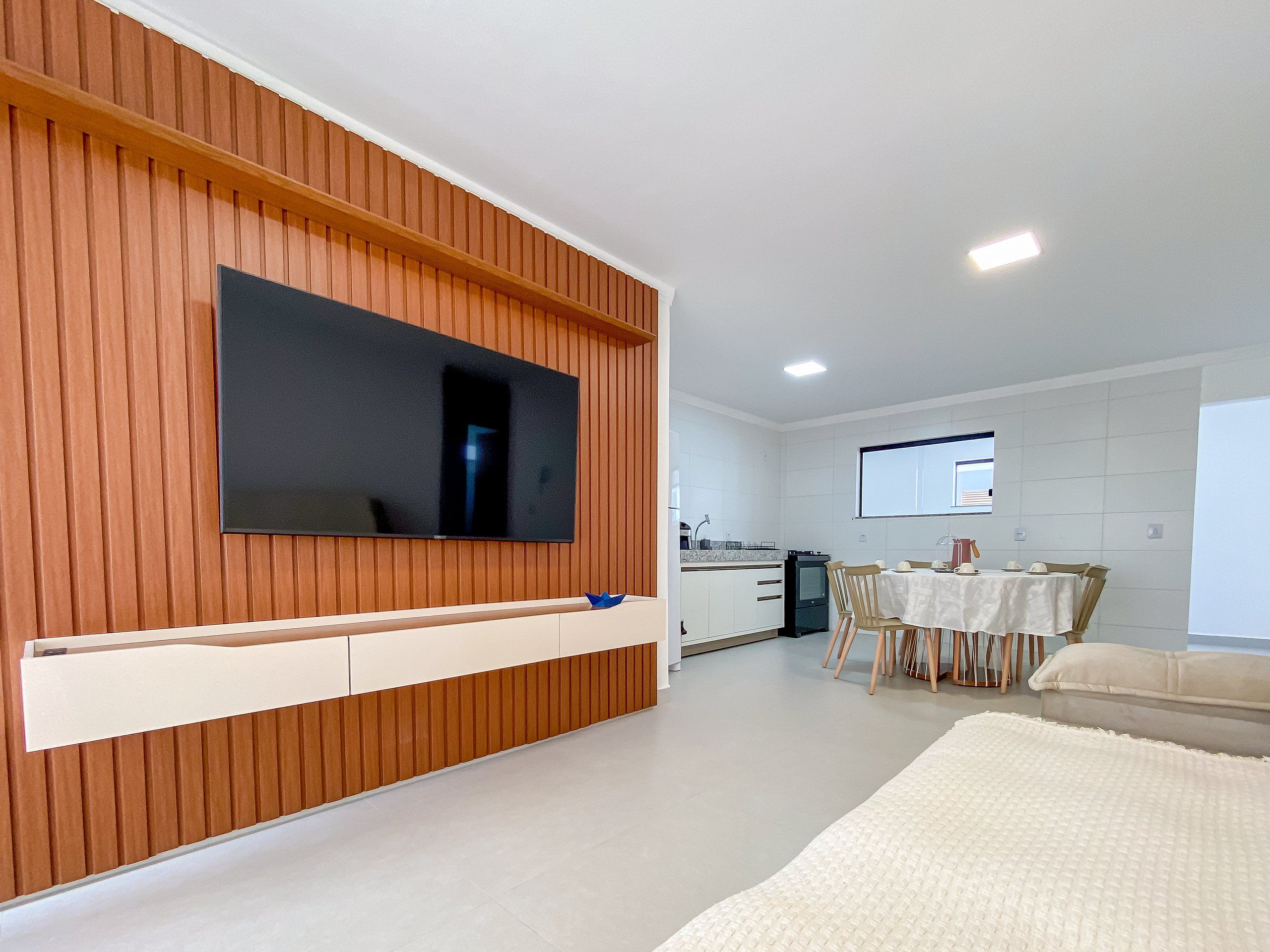 A modern living room with a flat-screen TV mounted on a wooden wall, a dining area with a table and chairs, and a kitchen in the background.