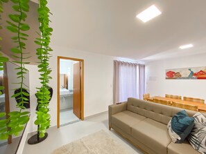 A living room with a sofa, a dining table with chairs, a hanging plant, and a door leading to a bathroom. - Residencial Saint Loius (Porto Seguro)