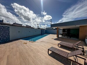 A rooftop swimming pool with a wooden deck, lounge chairs, and a covered seating area. - Residencial Saint Loius (Porto Seguro)