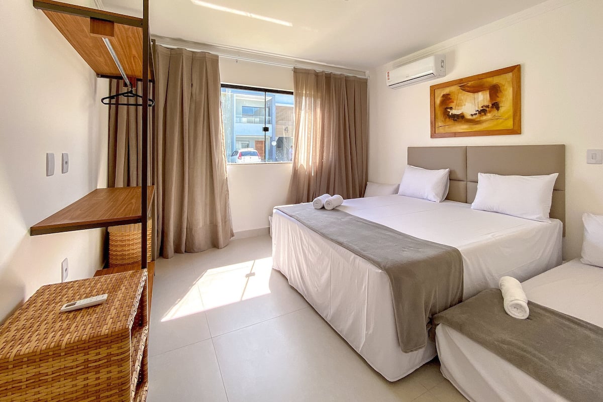 A hotel room with a bed, a bedside table, a window with curtains, an air conditioning unit, and a painting on the wall.