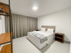 A hotel room with a bed, a nightstand, a wooden bench, and a large window with curtains. - Residencial Saint Loius (Porto Seguro)
