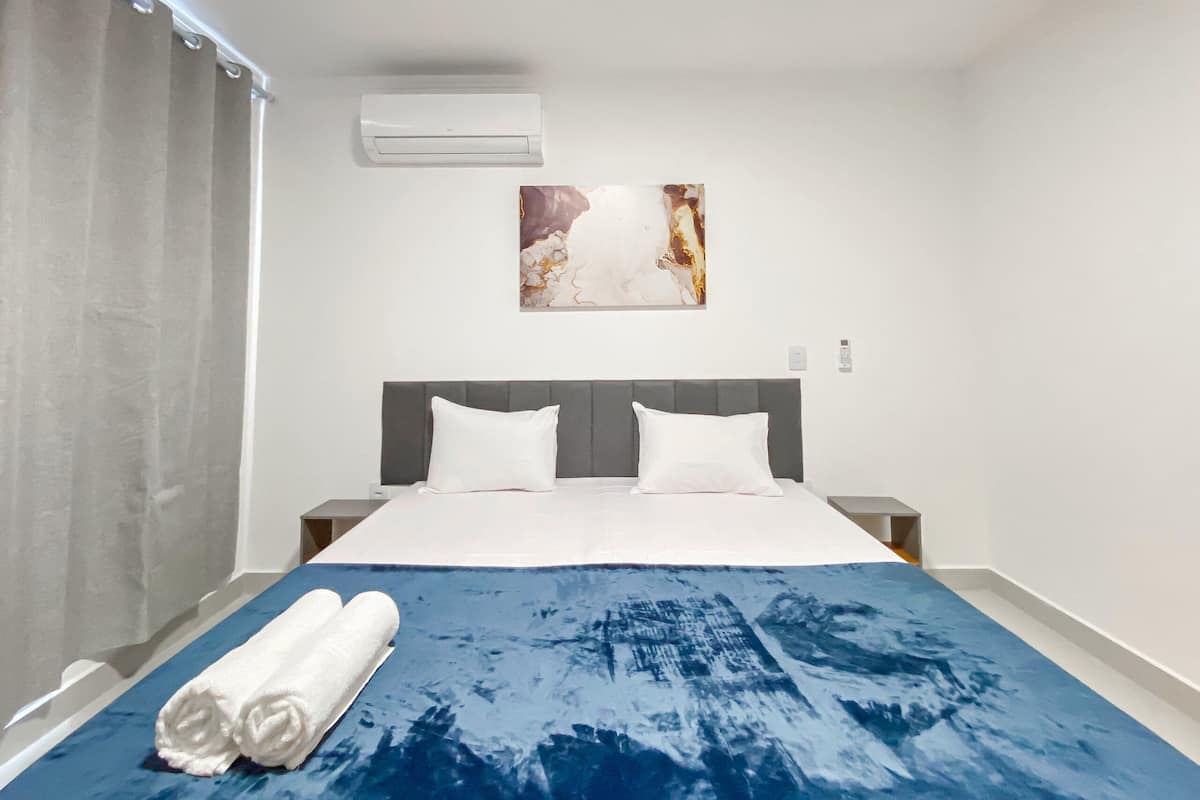 A hotel room with a bed, a painting on the wall, and an air conditioning unit.