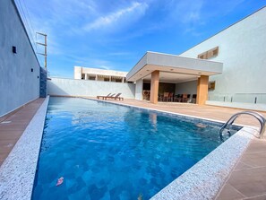 A swimming pool with a clear blue water, surrounded by a covered patio area with chairs and tables. - Residencial Saint Loius (Porto Seguro)