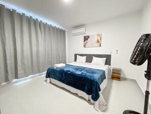 A bedroom with a bed, curtains, an air conditioner, a wall-mounted artwork, and a standing fan. - Residencial Saint Loius (Porto Seguro)