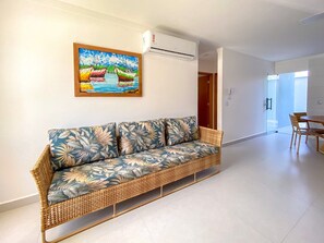 A wicker sofa with floral upholstery, a painting of boats on the wall, and a dining area with chairs and a table. - Residencial Saint Loius (Porto Seguro)