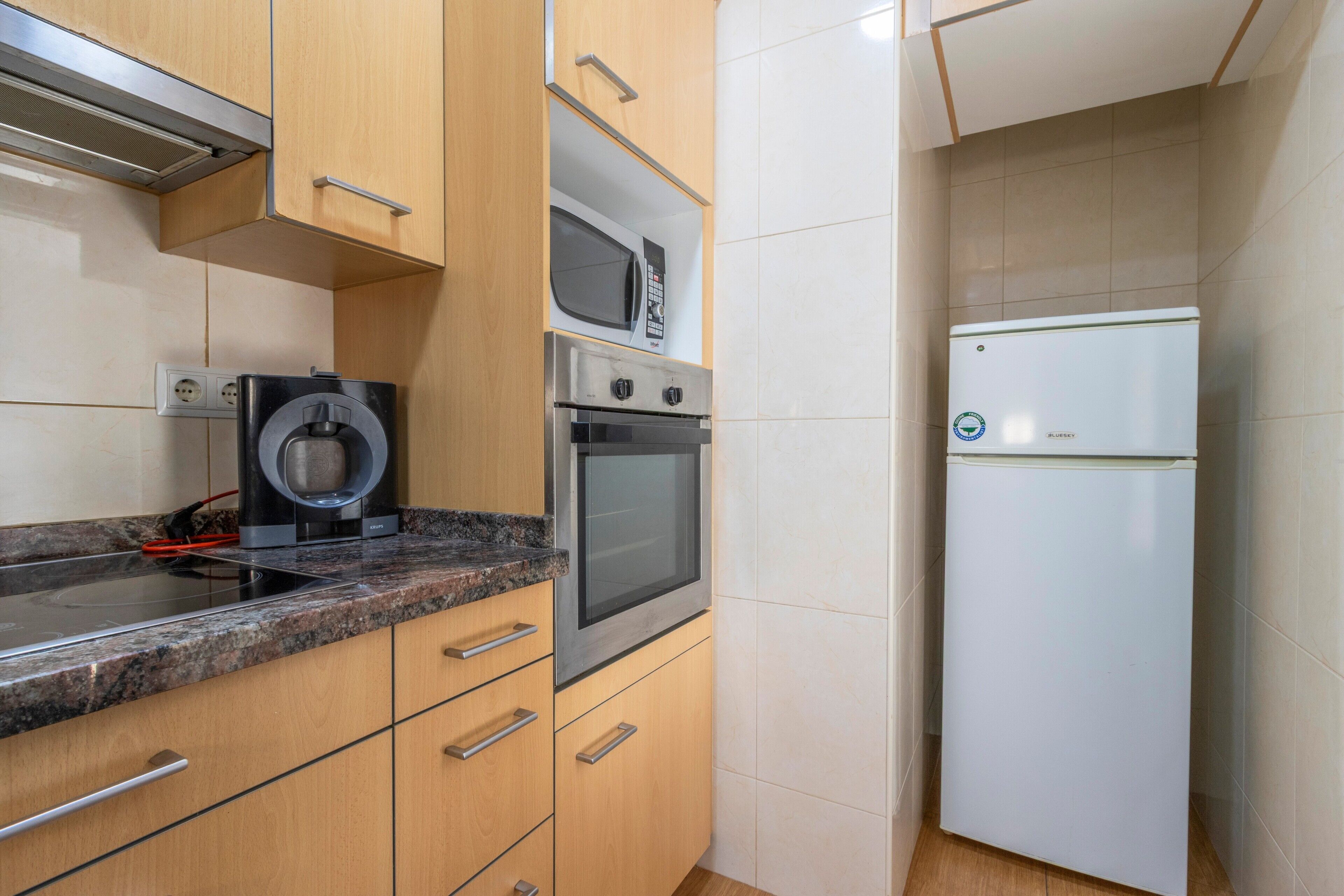 Una cocina compacta con microondas, horno y refrigerador integrados, con armarios de madera y encimera de granito.