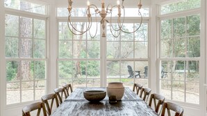 Une salle à manger avec une grande table en bois, des fenêtres à cadre blanc et un lustre.