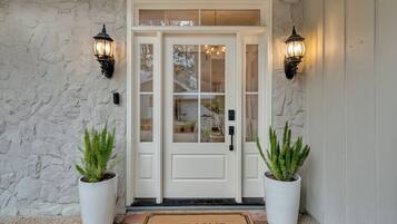 Une porte blanche avec des panneaux de verre, flanquée de deux plantes en pot et de deux lampes fixées au mur.