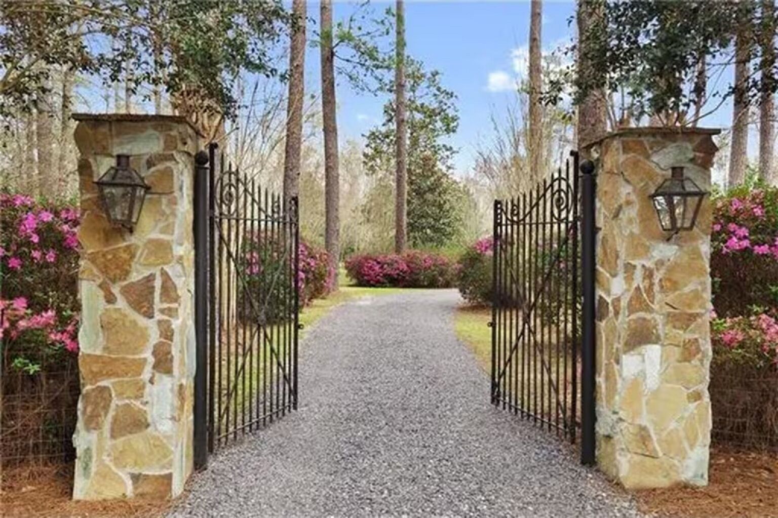 A gravel pathway leading through a gated entrance with stone pillars and lanterns, surrounded by trees and flowering shrubs.