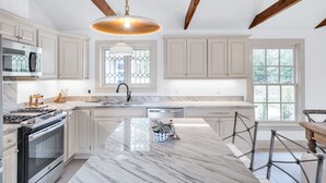 Une cuisine moderne avec des meubles blancs, un îlot en marbre, une cuisinière en acier inoxydable et une grande fenêtre donnant sur un paysage verdoyant.