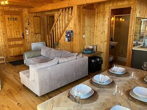 L’intérieur d’un chalet en bois chaleureux et accueillant, avec un canapé, une table à manger pour quatre personnes et un coin cuisine.
