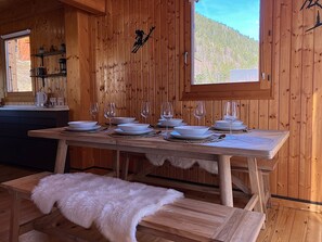 Interior d'una cabana de fusta amb una taula de dinar preparada per a sis, una banca amb una manta de pell, i una finestra amb vista a una muntanya boscosa.