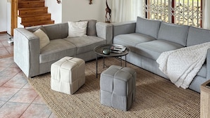 Una sala de estar amplia con un sofá seccional grande de color gris, un pequeño reposapiés y una mesa de centro. Se ve una escalera de madera al fondo, que conduce a otro nivel.