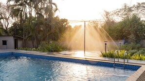 A swimming pool with a sunlit edge, surrounded by greenery and a small building. - Glory of MJ, Family Resort in Nature, Panvel (Panvel)