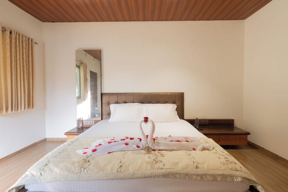 Un lit bien fait, recouvert d’une couvre-lit à motifs floraux, avec une tête de lit en bois et une table de chevet en bois sur laquelle se trouve une lampe.