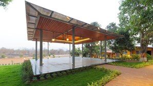 A covered outdoor area with a metal roof and columns, surrounded by greenery and a brick pathway. - Glory of MJ, Family Resort in Nature, Panvel (Panvel)