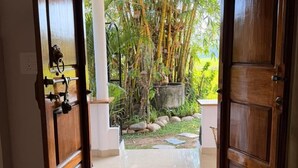 An open wooden door leading to a garden with bamboo, a stone planter, and a tiled floor. - Bhoomi by Ostello (Varkala)