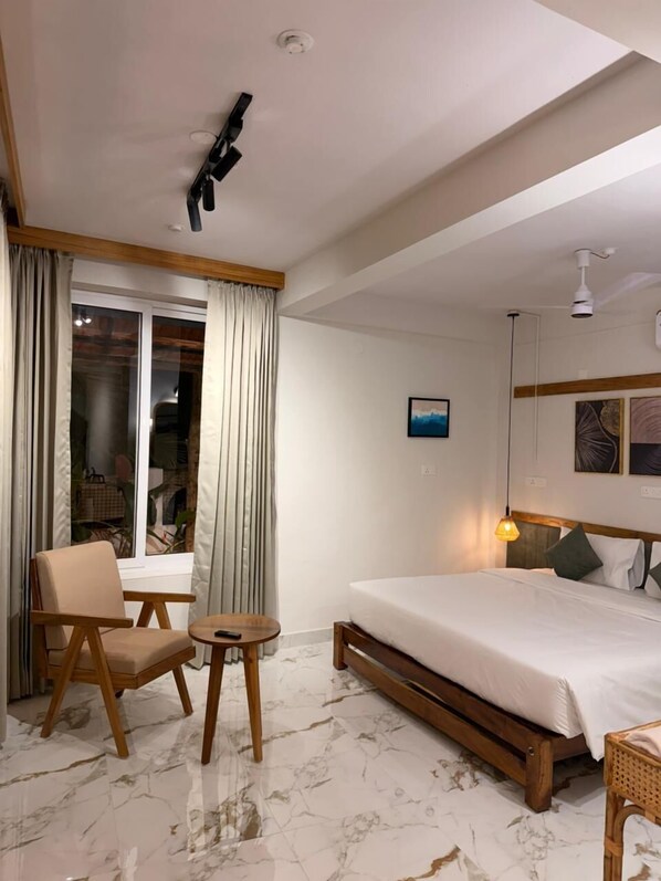 Habitación de hotel con cama, una silla, una mesita pequeña, ventilador de techo y ventana con cortinas.
