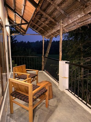 A wooden balcony with two chairs and a table, a thatched roof, and a view of trees. - Bhoomi by Ostello (Varkala)