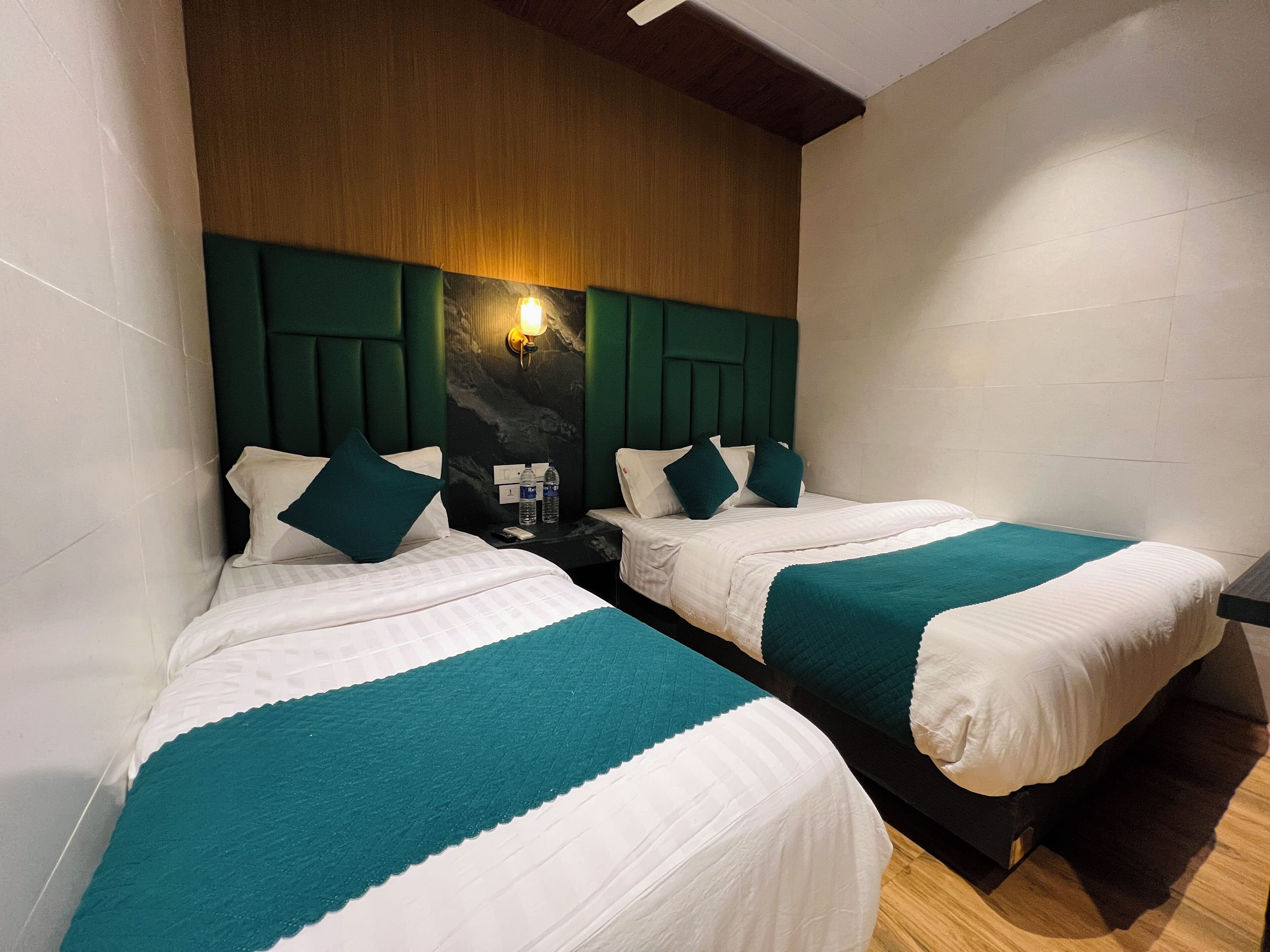 A hotel room with two beds, each with white bedding and teal accents, a wooden headboard, and a wall-mounted light fixture.