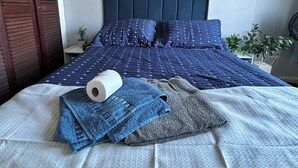 Un lit bien fait, avec une couette bleue, un oreiller blanc et une serviette bleue. On peut voir une petite plante sur la table de chevet.