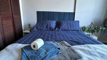 Cama bem arrumada com colcha azul, travesseiro branco e toalha azul. Há uma pequena planta na mesa de cabeceira.