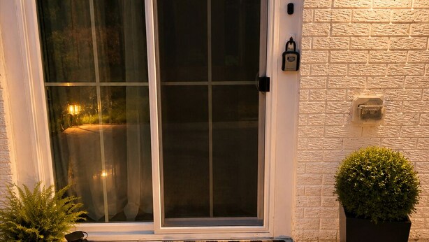 Une porte à cadre blanc avec un panneau de verre, un paillasson accueillant, une plante en pot, une lanterne et un luminaire fixé au mur.