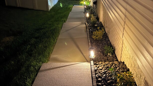 Un sendero que conduce a una casa con valla blanca y una fila de luces a lo largo del costado.