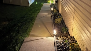 Un sendero que conduce a una casa con valla blanca y una fila de luces a lo largo del costado.