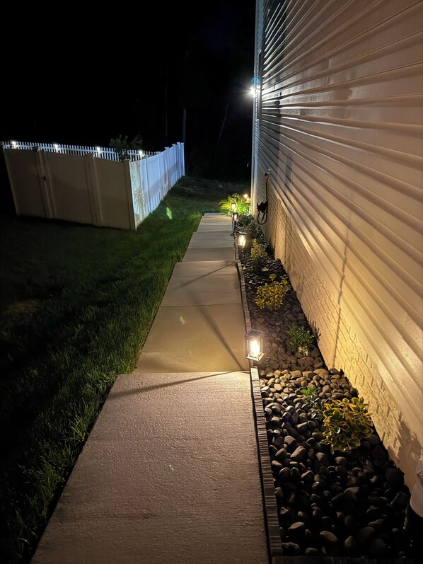 Un sendero que conduce a una casa con valla blanca y una fila de luces a lo largo del costado.