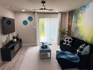 A living room with a flat-screen TV, a ceiling fan, a sofa with a blue throw blanket, and a large window with sheer curtains. - Modern Private Lowerlevel Suite 
Free Parking + Wi-Fi, close to Quantico  (Stafford)